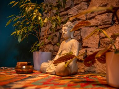 A stone Buddha statue seated on a colorful patterned rug with potted plants and a singing bowl at The Waterfall Retreat in Orton