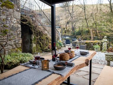 An outdoor patio dining area with a wooden table set with bread wine glasses and bowls near stone steps and garden at The Waterfall Retreat in Orton