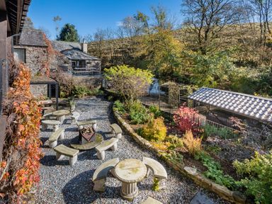 An outdoor garden area with stone benches tables a fire pit and a small waterfall near a stone house at The Waterfall Retreat in Orton