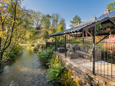 A covered outdoor seating area with couches next to a river surrounded by trees and plants at The Waterfall Retreat in Orton