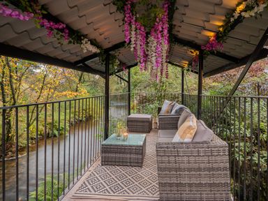 An outdoor covered patio with wicker sofa and coffee table overlooking a river at The Waterfall Retreat in Orton