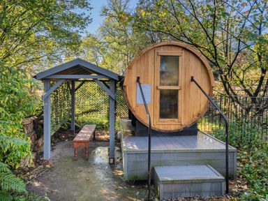 An outdoor wooden sauna barrel with steps and handrails next to a covered bench in a garden at The Waterfall Retreat in Orton
