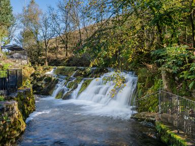 A waterfall flowing over rocks surrounded by trees and greenery with a seating area on the left at The Waterfall Retreat in Orton