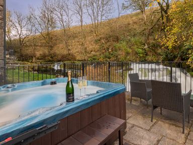 An outdoor patio with a hot tub champagne bottle and two glasses with chairs and a waterfall in the background at The Waterfall Retreat in Orton