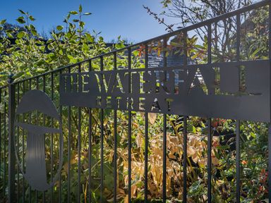 A metal gate with the words The Waterfall Retreat and a mushroom design surrounded by plants at The Waterfall Retreat in Orton
