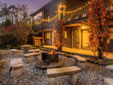 An outdoor seating area with curved stone benches around a fire pit outside a stone building with string lights and climbing plants at The Waterfall Retreat in Orton