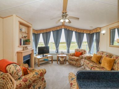 A living room with floral sofas a television on a wooden table blue curtains and a ceiling fan at Cae Gog Near Cemaes Bay