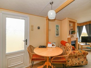 A dining area with a wooden round table and two wicker chairs next to a door and a living room with a floral sofa a fireplace and a television at Cae Gog Near Cemaes Bay