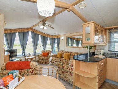 A living room with floral sofas and blue curtains next to a kitchen area with wooden cabinets at Cae Gog Near Cemaes Bay