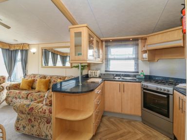 A kitchen with wooden cabinets stove and sink next to a living room with floral sofas and curtains at Cae Gog Near Cemaes Bay