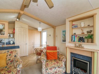 A living room with floral armchairs a fireplace shelving unit with books and decor and a dining area with a table and chairs at Cae Gog Near Cemaes Bay
