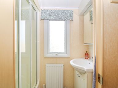 A bathroom with a sink cabinet a window with a patterned valance a radiator and a shower enclosure at Cae Gog Near Cemaes Bay