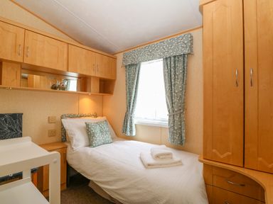 A bedroom with a single bed white bedding folded towels wooden cabinets and a window with patterned curtains at Cae Gog Near Cemaes Bay