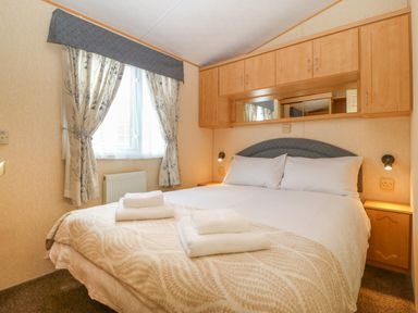 A bedroom with a double bed towels on the bed built in wooden cupboards above bed and a window with patterned curtains at Cae Gog Near Cemaes Bay