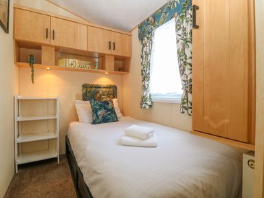 A small bedroom with a single bed white bedding and towels wooden cupboards and tropical patterned curtains at Cae Gog Near Cemaes Bay
