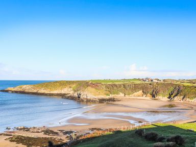 A coastal bay with sandy beach and rocky cliffs with grassy fields and buildings in the background at Cae Gog Near Cemaes Bay