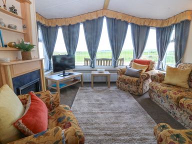 A living room with floral sofas a carpet a television on a stand and large windows with blue curtains at Cae Carreg in Cemaes Bay