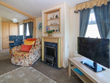 A living room with a floral armchair near a fireplace and a television on a wooden stand at Cae Carreg in Cemaes Bay
