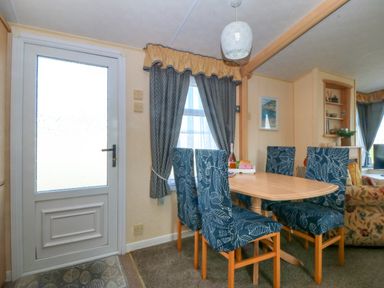 A dining area with a wooden table and six blue patterned chairs near a door and windows with curtains at Cae Carreg in Cemaes Bay