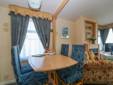 A dining area with a wooden table and four chairs with patterned covers next to a window with curtains and a living room with a floral sofa and shelves at Cae Carreg in Cemaes Bay