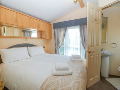 A bedroom with a double bed, white linens, towels on the bed, wooden cabinets above the bed, and an open door showing a sink and mirror at Cae Carreg in Cemaes Bay
