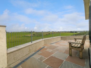 A stone tiled patio with a wooden bench and metal railing overlooking green fields at Cae Carreg in Cemaes Bay