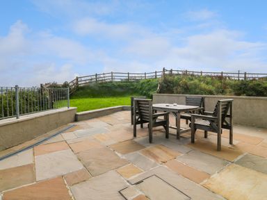 An outdoor patio with a wooden table and four wooden chairs surrounded by a low wall and grassy area at Cae Carreg in Cemaes Bay