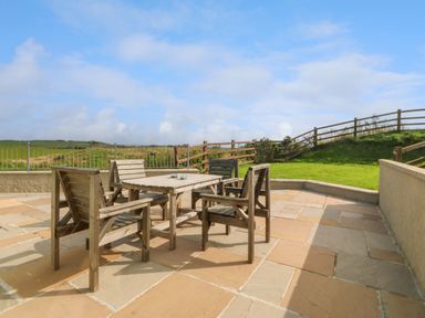 An outdoor patio with a wooden table and four chairs on stone tiles overlooking a grassy area with wooden fences at Cae Carreg in Cemaes Bay