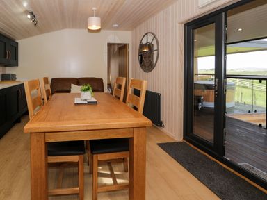A dining area with a wooden table and six chairs next to black framed glass doors opening to an outdoor deck with a hot tub at Betsi Lodge 1 Llanelidan near Corwen