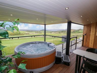 A covered outdoor patio with a hot tub table chairs and steps overlooking a grassy field with hills in the distance at Betsi Lodge 1 Llanelidan near Corwen