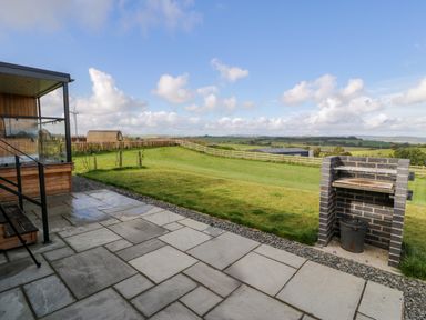 An outdoor patio area with tiled flooring a brick barbecue and a grassy lawn with a wooden fence and countryside views at Betsi Lodge 1 in Llanelidan near Corwen