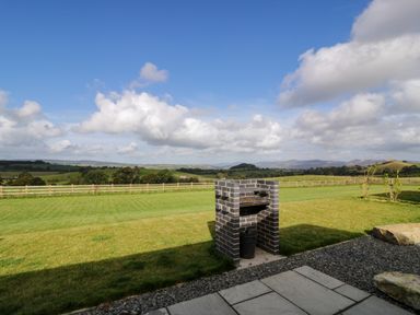 An outdoor brick barbecue on a grassy lawn with a wooden fence and hills in the background at Betsi Lodge 1 in Llanelidan near Corwen