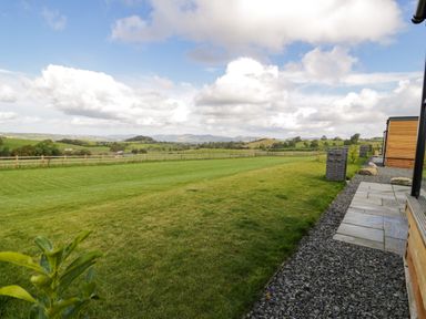 A grassy yard with a stone path and a wooden building overlooking rolling hills and a cloudy sky at Betsi Lodge 1 in Llanelidan near Corwen