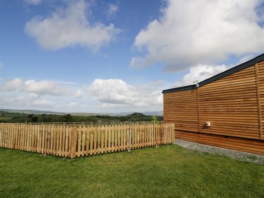 A fenced garden area next to a wooden building with grassy fields and hills in the background at Betsi Lodge 1 in Llanelidan near Corwen