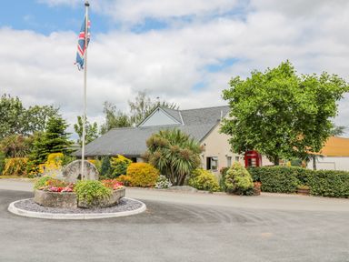 A roundabout with flowers and a flagpole in front of a house with bushes and trees at Hazel Lodge in Teigngrace near Newton Abbot