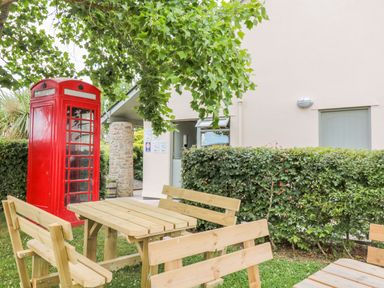 An outdoor seating area with wooden picnic tables and benches near a red telephone booth and a building at Hazel Lodge in Teigngrace near Newton Abbot