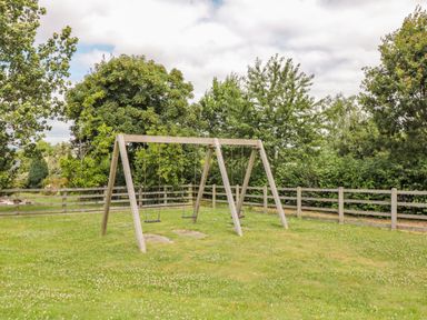 A wooden swing set on a grassy area surrounded by a wooden fence and trees at Hazel Lodge in Teigngrace near Newton Abbot