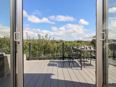 A deck with outdoor table and chairs set for tea with glass railing and hedge in the background at Hazel Lodge in Teigngrace near Newton Abbot