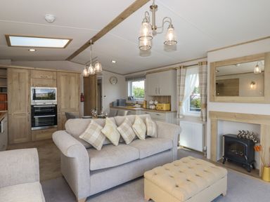 A living room with a sofa, an ottoman, a kitchen area with wooden cabinets, and a wall-mounted electric fireplace at Hazel Lodge in Teigngrace near Newton Abbot