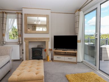 A living room with a TV on a wooden stand a fireplace with a mirror above and sliding glass doors to a deck at Hazel Lodge in Teigngrace near Newton Abbot