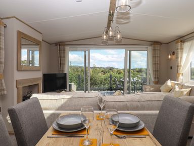 A dining table set with plates and glasses in front of a living room with sofas and sliding glass doors leading to an outdoor seating area with a view of trees at Hazel Lodge in Teigngrace near Newton Abbot