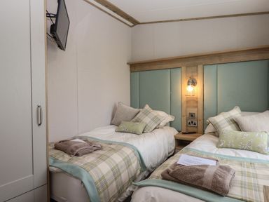 A bedroom with two single beds with plaid bed runners and pillows a wall-mounted TV and a bedside table with a lamp at Hazel Lodge in Teigngrace near Newton Abbot
