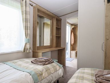 A bedroom with two single beds with folded towels on them a wooden vanity with a mirror a window with curtains and an open door showing a coat hanging at Hazel Lodge in Teigngrace near Newton Abbot