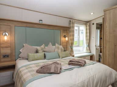 A bedroom with a bed with multiple pillows and folded towels window with curtains wooden furniture and a door leading to a bathroom at Hazel Lodge in Teigngrace near Newton Abbot