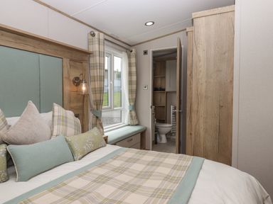 A bedroom with a bed, pillows, window with curtains, bench seat, and a door leading to a bathroom at Hazel Lodge in Teigngrace near Newton Abbot