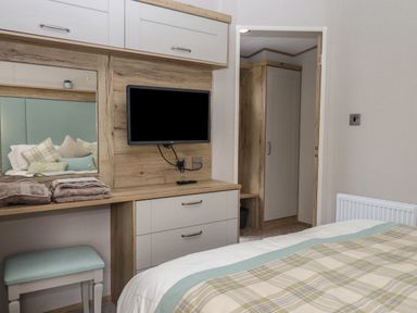 A bedroom with a bed plaid bedding a TV on a wooden dresser and a vanity with a bench at Hazel Lodge in Teigngrace near Newton Abbot