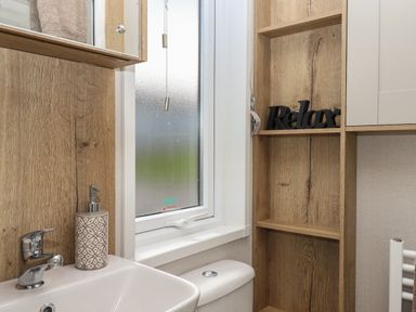 A bathroom with a white sink and toilet a window with a blind and wooden shelving with a relax sign at Hazel Lodge in Teigngrace near Newton Abbot