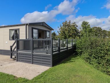 A mobile home with a deck and outdoor furniture next to a hedge under a blue sky at Hazel Lodge in Teigngrace near Newton Abbot