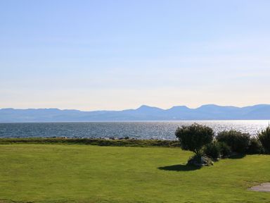 A grassy lawn with bushes by a body of water with mountains in the background at Seafront Holiday Home 2 in Pwllheli
