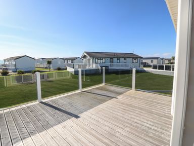 A wooden deck with glass railings overlooking a grassy area and several white holiday homes at Seafront Holiday Home 2 in Pwllheli